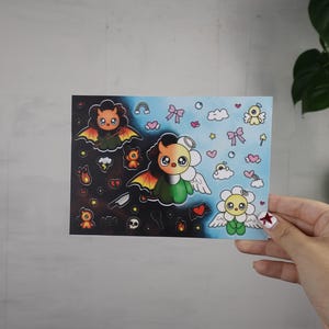 Może przedstawiać: Arkusz naklejek z podzielonym wzorem przedstawiającym urocze, kreskówkowe postacie. Jedna strona jest ciemna z nietoperzami i płomieniami, druga jasnoniebieska z aniołami i kokardkami. Centralna postać to pół diabeł, pół anioł.