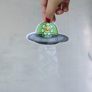 May include: A green and yellow cartoon flower with a sad face is inside a gray flying saucer. The sticker is on a clear plastic sheet.