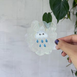 May include: A clear, iridescent sticker in the shape of a cloud with a cute cloud and raindrops design. The sticker is held by a hand against a light gray background with green leaves.
