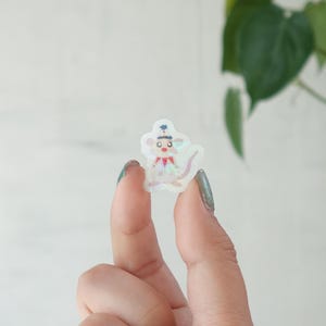 Puede incluir: Una pequeña pegatina holográfica con un ratón de dibujos animados con sombrero y pajarita. La pegatina tiene un brillo nacarado y se sostiene entre dos dedos sobre un fondo borroso de hojas verdes y una pared blanca.