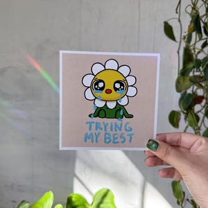 May include: A cartoon illustration of a yellow daisy with a green stem and leaves, crying with blue tears. The daisy is sitting on a light pink background with the text "TRYING MY BEST" written in blue.
