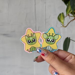 May include: Two stickers featuring cartoon alien-like creatures inside yellow star shapes. The creatures have large eyes and are colored green, with a pink and blue outline. The stickers are held by a hand with colorful nail art.