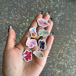 May include: A collection of colorful, small stickers featuring various cartoon animal designs. The stickers include a pink dinosaur, a red balloon dog, a black cat, and other whimsical characters. The stickers are held in a hand against a sparkly background.