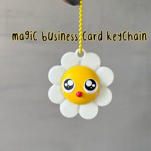 May include: A white daisy-shaped keychain with a yellow center and a cartoon face. The face has black eyes, a red nose, and a small smile. The keychain is attached to a gold chain.