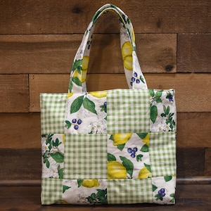 May include: A patchwork tote bag with a green and white gingham pattern, and panels featuring lemons, blueberries, and floral designs. The bag has matching handles and is set against a wooden background.