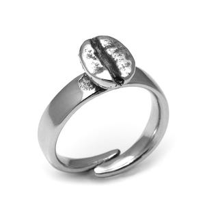 Puede incluir: Anillo plateado con un diseño detallado de grano de café. El anillo tiene una banda ajustable y un grano de café detallado. El anillo se presenta sobre un fondo blanco.