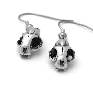 May include: A pair of silver-colored earrings featuring small, detailed skull pendants. The skull pendants have visible eye sockets and nasal cavities, and are suspended from simple hook-style ear wires. The earrings are displayed against a white background.