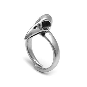 Op de afbeelding: Een zilverkleurige ring met een vogel schedel ontwerp. De schedel heeft een puntige snavel en een holle oogkas. De ringband is glad en verstelbaar, met een eenvoudig, elegant ontwerp.
