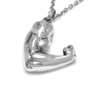 May include: A silver-colored pendant necklace featuring a sculpted arm flexing a bicep. The pendant is attached to a delicate chain. The jewelry is made of a shiny, metallic material and is set against a white background.