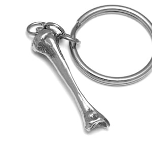 Puede incluir: Un llavero de plata con una réplica detallada de un hueso humano. El hueso está unido a un anillo de llave de plata.
