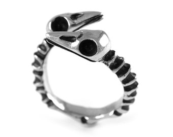 Puede incluir: Un anillo de plata con un diseño esquelético que presenta dos cráneos de pájaros. El anillo está hecho de metal texturizado y tiene un diseño detallado.