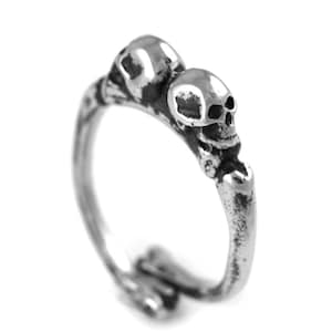 Puede incluir: Un anillo de plata con dos diseños de calavera. Las calaveras están mirando hacia adelante y están conectadas por una banda ósea.