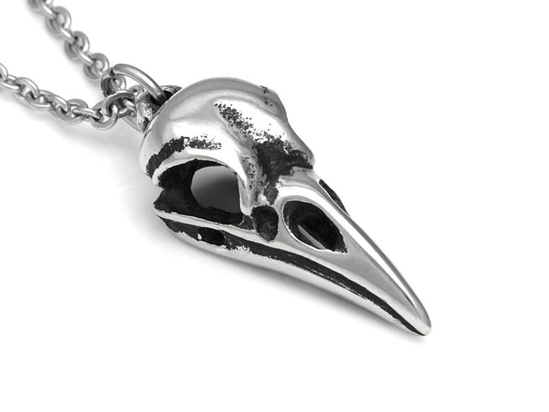 May include: A silver pendant necklace featuring a detailed crow skull design. The pendant is made of metal and has a textured finish.
