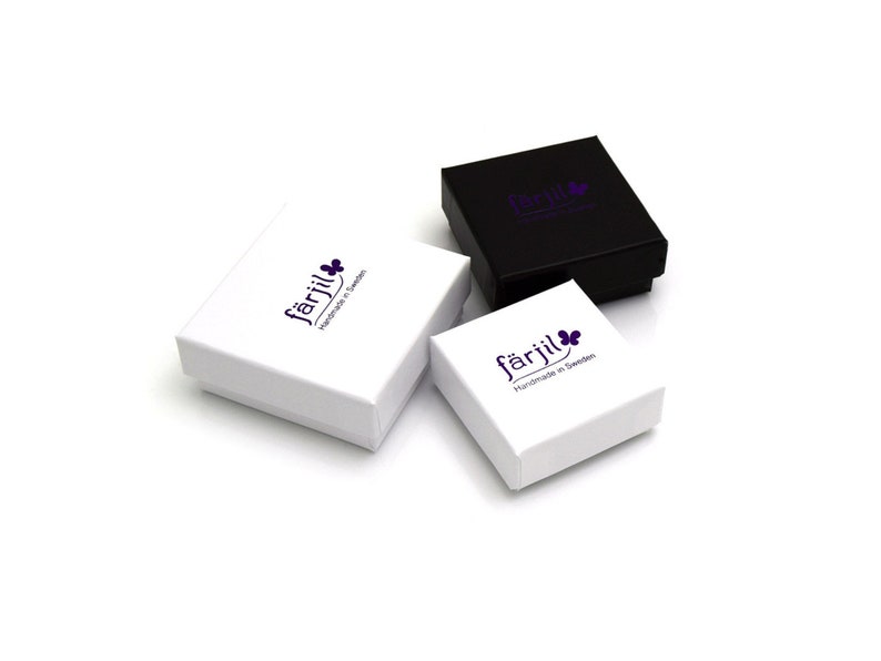 May include: Three small jewelry boxes, two white and one black, are arranged on a white surface. The boxes have a square shape with a lid. Each box features the word "f&auml;rjil" in a purple font, along with a purple flower graphic and the text "Handmade in Sweden."