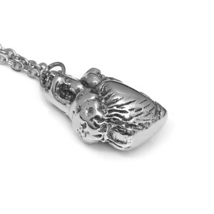 May include: Silver-toned boxing glove pendant necklace. The pendant is detailed with textured surfaces, and is attached to a delicate chain. The necklace is displayed against a white background, highlighting the metallic sheen of the jewelry.