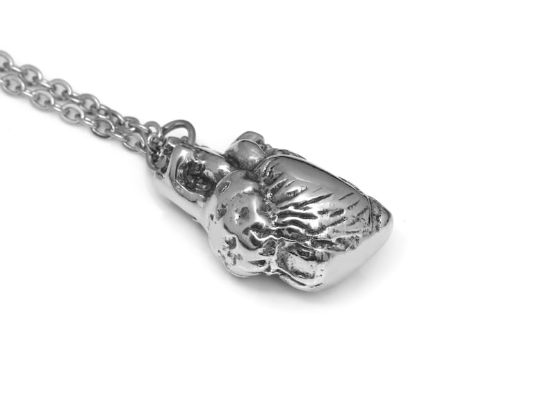 May include: Silver-toned boxing glove pendant necklace. The pendant is detailed with textured surfaces, and is attached to a delicate chain. The necklace is displayed against a white background, highlighting the metallic sheen of the jewelry.