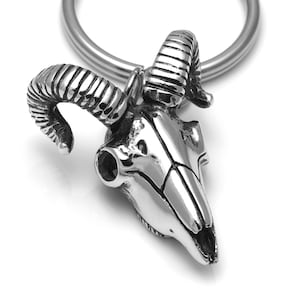 May include: A silver-toned keychain featuring a detailed ram skull with curled horns. The keychain is attached to a silver-toned ring.