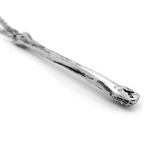 May include: A silver necklace with a long, thin pendant that resembles a branch or twig. The pendant has a rough, textured surface.