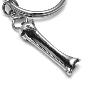 May include: A silver keychain featuring a detailed bone charm. The bone charm is a realistic representation of a human bone, with a smooth, polished finish.