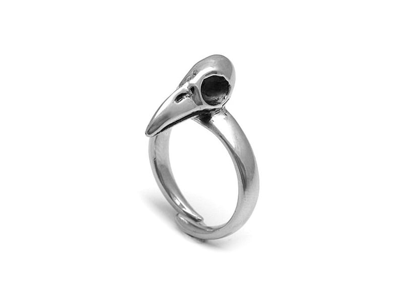 May include: A silver-colored ring featuring a bird skull design. The skull has a pointed beak and a hollow eye socket. The ring band is smooth and adjustable, with a simple, elegant design.