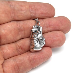May include: A silver-colored anatomical heart pendant on a chain. The pendant is detailed with veins and chambers, and is held in a hand against a white background. The pendant is a unique piece of jewelry.