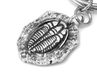 May include: A silver-colored keychain featuring a detailed trilobite fossil design. The fossil is set within a textured, irregular-shaped pendant, attached to a split ring. The keychain is made of metal and is suitable for everyday use.