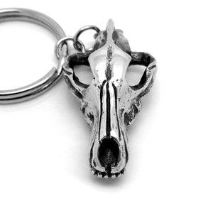 Peut inclure: Un porte-clés en métal argenté avec un charme en forme de crâne de loup détaillé. Le crâne est sculpté de manière réaliste avec une surface texturée et une mâchoire proéminente.