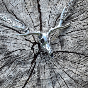 May include: A silver deer skull pendant necklace with a detailed design. The pendant is hanging on a silver chain and is resting on a wooden surface.
