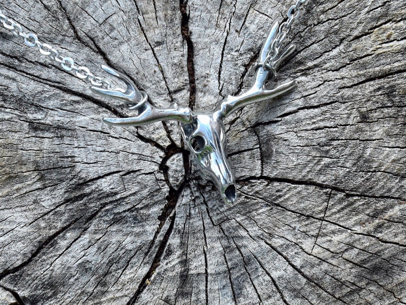 May include: A silver deer skull pendant necklace with a detailed design. The pendant is hanging on a silver chain and is resting on a wooden surface.