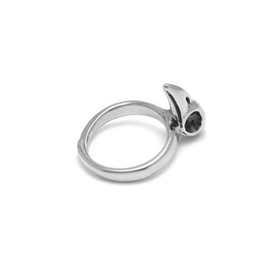 May include: A silver-toned ring with a bird skull design. The ring features a rounded band and a detailed skull at the top, with a dark, possibly black, stone set in the eye socket. The ring is set against a white background.