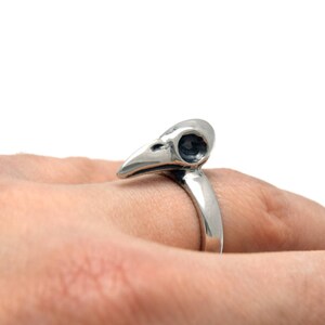 May include: A silver-colored ring featuring a bird skull design. The ring's band curves up to form the skull, with detailed eye sockets and beak. The ring is worn on a finger, showcasing the gothic and unique jewelry piece.