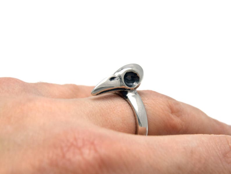 May include: A silver-colored ring featuring a bird skull design. The ring's band curves up to form the skull, with detailed eye sockets and beak. The ring is worn on a finger, showcasing the gothic and unique jewelry piece.