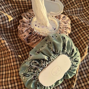 May include: Two fabric-covered wipe dispensers featuring a gathered, ruffled design. One dispenser is tan with a black paisley pattern, and the other is sage green with a black paisley pattern. Both have white plastic lids with a butterfly logo.