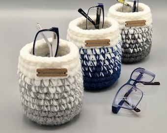 May include: Three cylindrical crochet holders with white rims and ombre bases in gray, blue, and dark gray. Each holder features a small wooden tag labeled OlgaAndrewDesigns. Two holders contain eyeglasses, and one pair of blue glasses rests nearby.