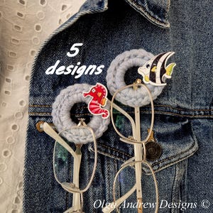 May include: Two light blue crocheted rings serving as eyeglass holders, each featuring a small decorative sea creature charm. One charm is a red seahorse, the other is a black and white angelfish. The text 5 designs is visible.