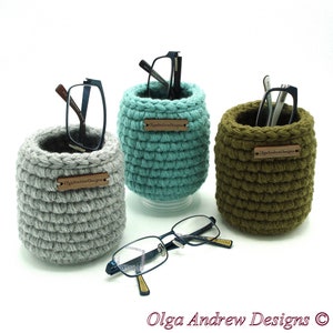Puede incluir: Tres contenedores cilíndricos de crochet gris, azul verdoso y verde oliva con una etiqueta marrón que dice "Olga Andrew Designs" en cada uno. Los contenedores están llenos de gafas.