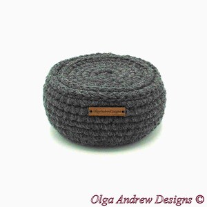 May include: A round, gray crocheted basket with a textured surface. The basket has a small, rectangular wooden tag that reads "OlgaAndrewDesigns". The basket is a handmade item.