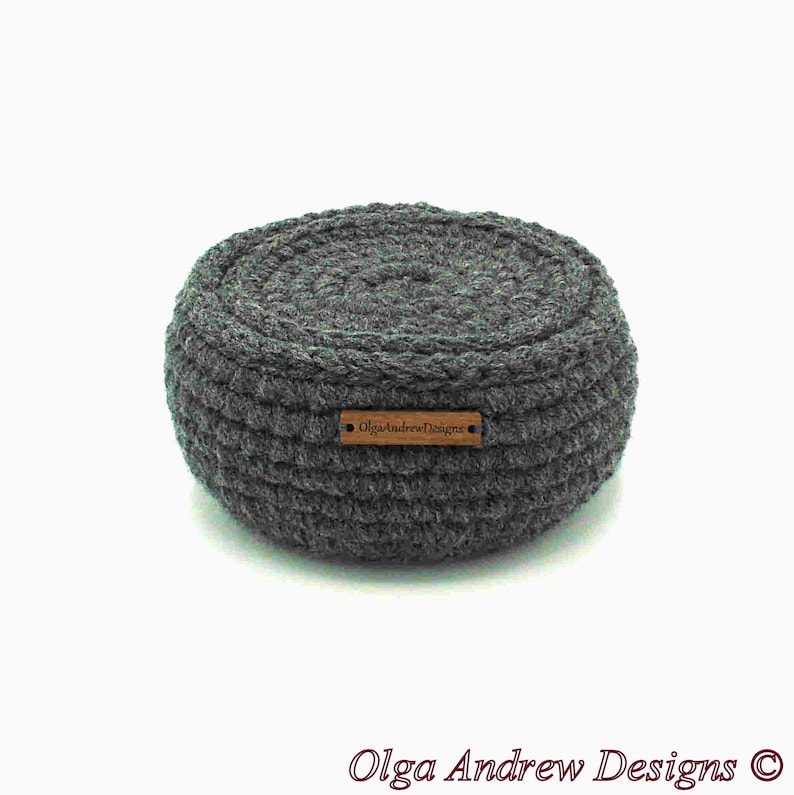 May include: A round, gray crocheted basket with a textured surface. The basket has a small, rectangular wooden tag that reads "OlgaAndrewDesigns". The basket is a handmade item.