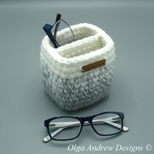 Puede incluir: Un portagafas de crochet blanco y gris con dos compartimentos. El portagafas tiene una etiqueta marrón que dice "Olga Andrew Designs".