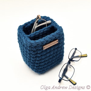 May include: A deep blue, square-shaped crochet basket with a textured stitch pattern. A small wooden tag labeled OlgaAndrewDesigns is attached to the side. A pair of glasses rests inside the basket, and another pair lies on the surface nearby.