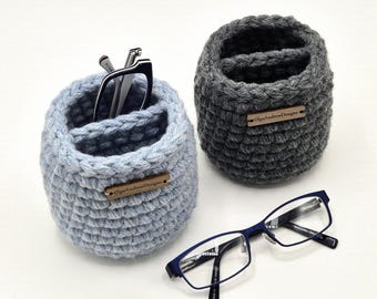 Puede incluir: Dos organizadores cilíndricos de ganchillo, uno azul claro y otro gris oscuro, cada uno con una pequeña etiqueta rectangular de madera con la inscripción OlgaAndrewDesigns. El organizador azul claro contiene unas gafas de montura negra, mientras que otras gafas de montura azul descansan al lado.