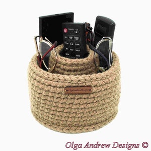 May include: A beige, hand-crocheted organizer basket with two compartments. The basket holds a remote control, glasses, and a small black item. A small wooden tag with the text "Olga Andrew Designs" is attached to the front of the basket.