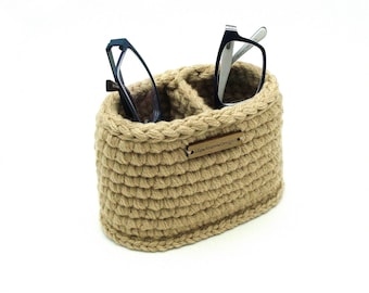 Puede incluir: Un organizador ovalado de ganchillo en color beige con dos compartimentos internos, que contiene dos pares de gafas. Una pequeña etiqueta rectangular de madera está fijada en la parte frontal con el texto grabado OlgaAndrewDesigns.