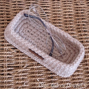 May include: A beige crocheted eyeglass case with a pair of silver-rimmed glasses inside. The case is sitting on a woven wicker surface.