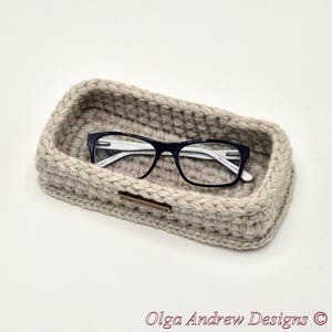 May include: A beige, rectangular crochet basket with a pair of black-framed eyeglasses. The basket has a thick, textured appearance and a small, rectangular tag. The text "Olga Andrew Designs" is at the bottom.