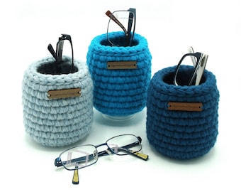 May include: Three cylindrical crochet holders in light blue, bright blue, and dark blue, each featuring a small brown rectangular tag labeled OlgaAndrewDesigns. Two holders contain eyeglasses, and a pair of glasses rests in front on the surface.
