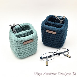 May include: Two handmade crochet eyeglass holders in teal and navy blue, each with two compartments. The teal holder has black-framed glasses, and the navy blue holder has gold-framed glasses. The text "Olga Andrew Designs" is at the bottom.