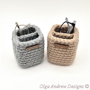May include: Two square crochet organizers, one light gray and one beige, each featuring a small wooden tag labeled OlgaAndrewDesigns. The gray organizer holds a pair of silver-rimmed glasses, while the beige organizer holds black-rimmed glasses.