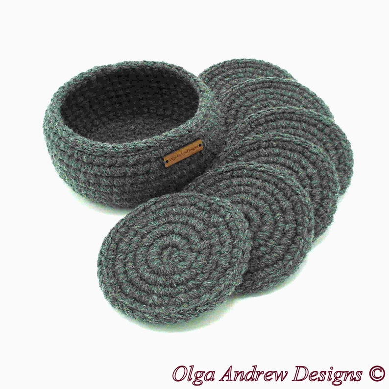 May include: A set of gray crocheted coasters and a matching basket. The coasters are round and arranged next to the basket. The basket has a small, rectangular tag. The text "Olga Andrew Designs" is at the bottom.