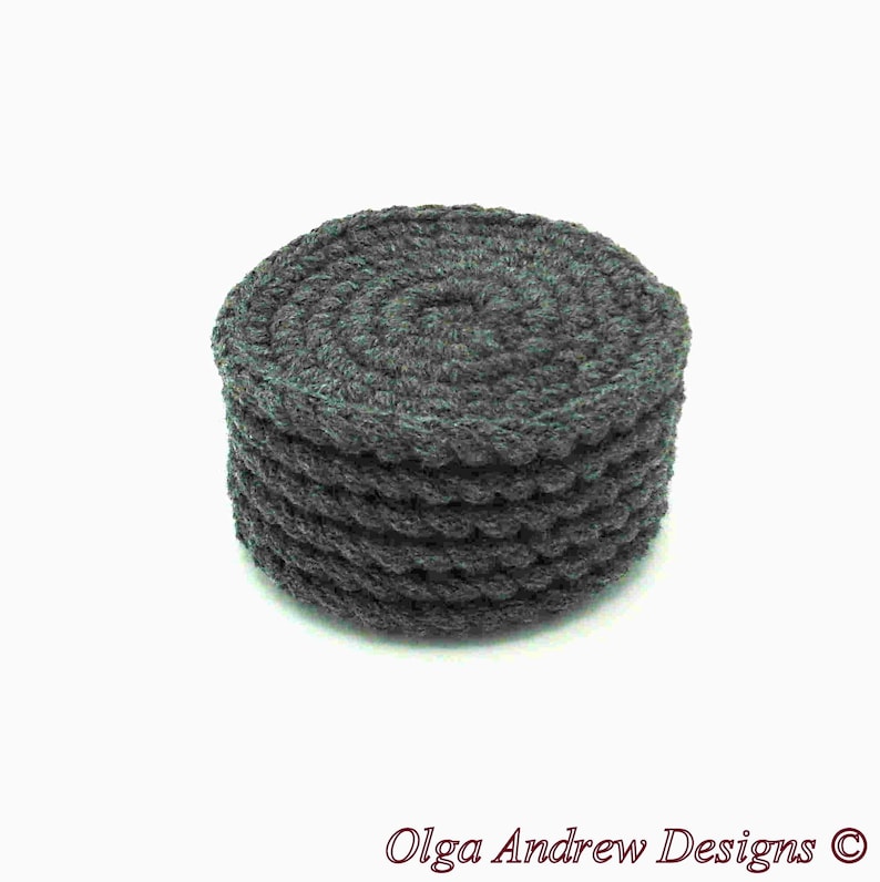May include: A dark gray, round, crocheted coaster. The coaster is made of thick yarn and has a textured, spiral design on top. The sides have a scalloped edge. The text "Olga Andrew Designs" is at the bottom.